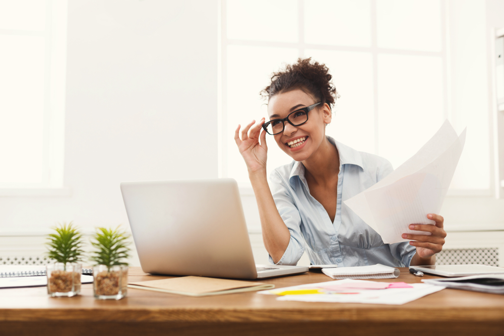 A smiling woman in glasses at a desk looking at her laptop with papers in her hand.