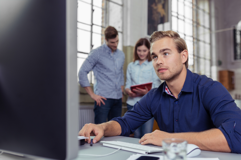 A man working on a computer with another couple in the background.