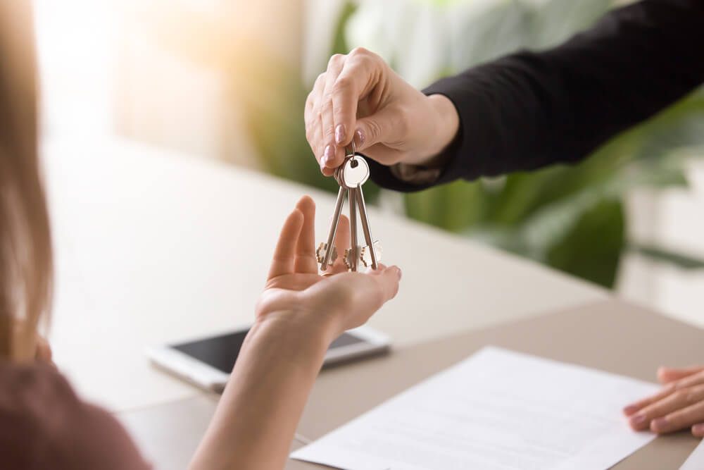 A person handing another person a set of keys over a desk with paper and a tablet.