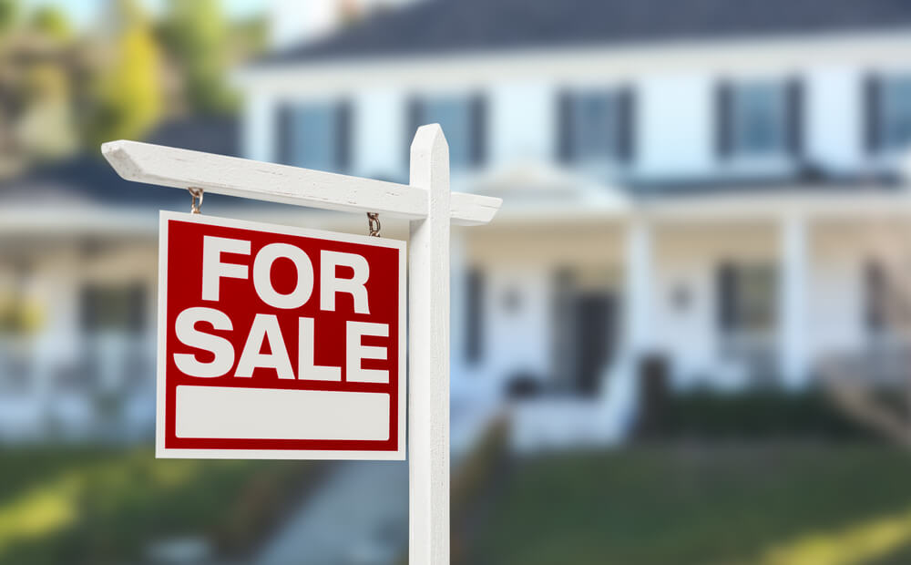 A for sale sign in front of white real estate investing property that is blurred out.