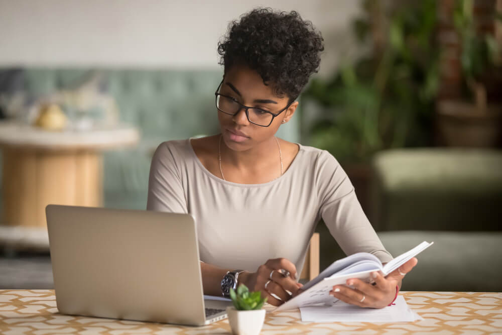 A woman in glasses studying a laptop with a booklet in her hands.