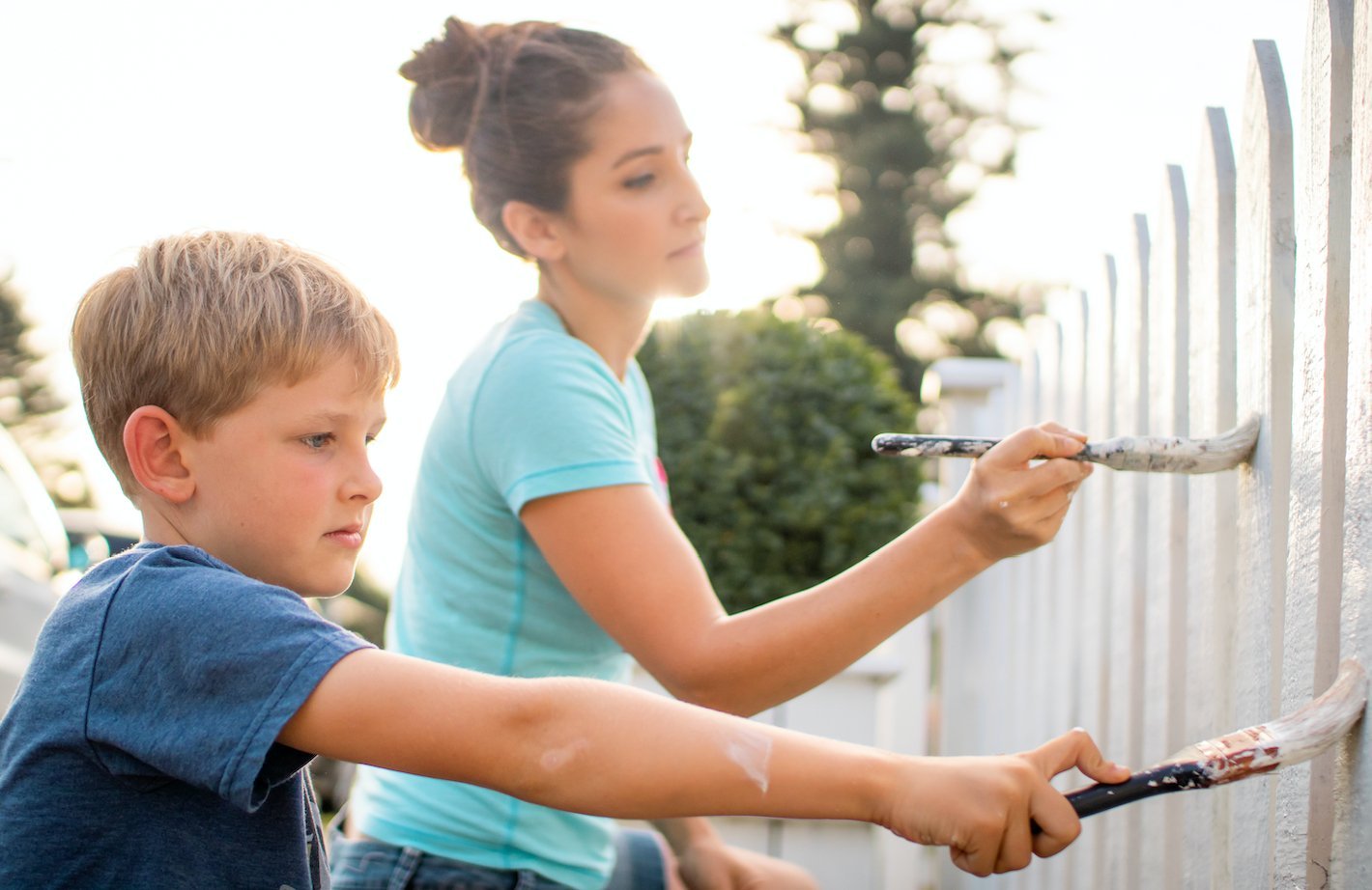 A young woman and small boy painting a white picket fence together.