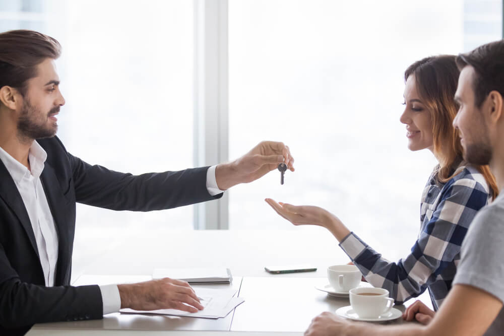 A man handing over a set of keys to couple at a desk.