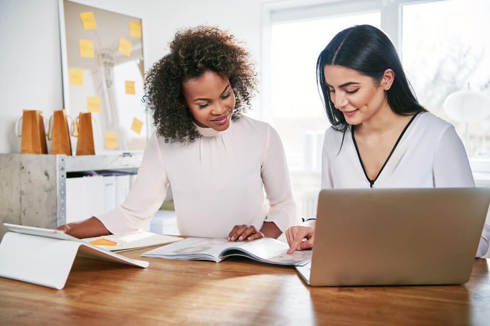 Two women looking over a booklet at a desk in front of a laptop.