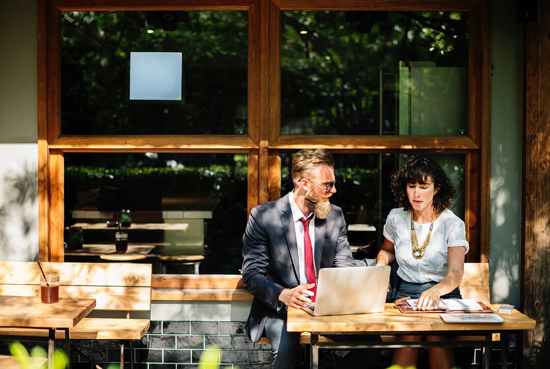 A man and woman at a small table outside a cafe in front of a laptop.