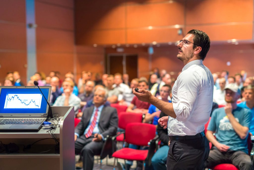 A keynote speaker in front of an audience with a microphone looking up at a presentation.