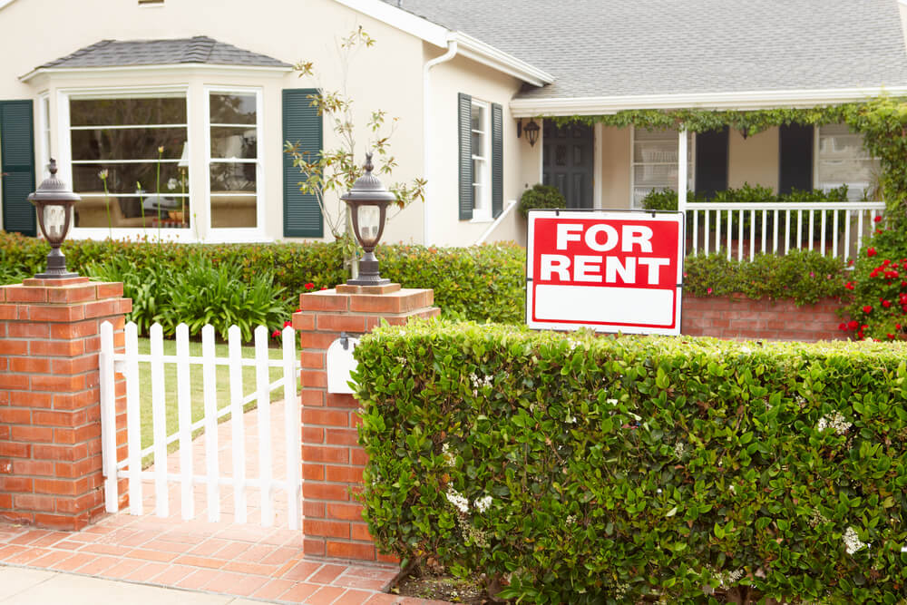 A white house with a white fence and green hedges with a red for sale sign.