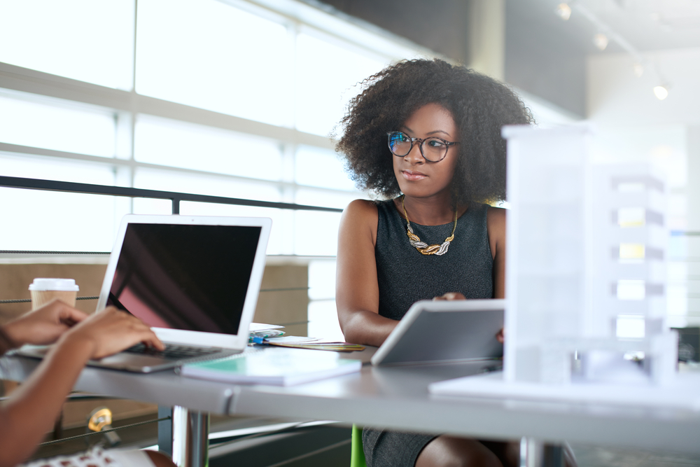 A woman on a laptop at a desk looking at another person on a laptop.