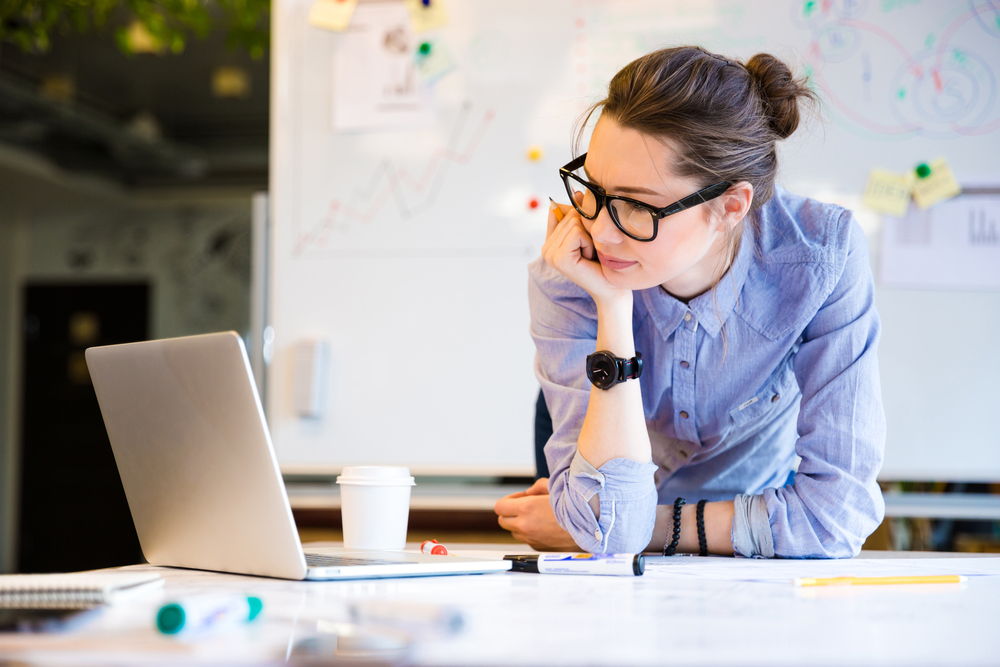 A woman with a white board behind her reaching a real estate investing business plan on a lap top.