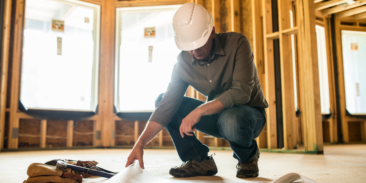 A construction manager reviews blueprints on a building site inside a structure with exposed wooden beams and sunlight streaming through windows.