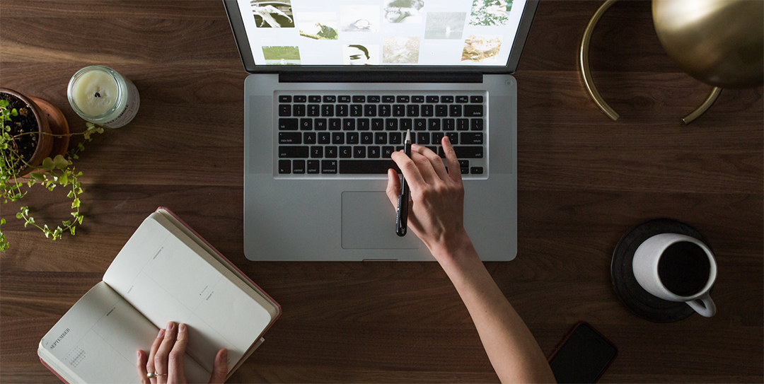 A woman's hand holding a pen and typing at laptop on her desk while looking at notes.